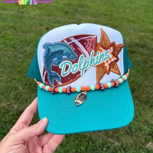 May include: A teal and white trucker hat with a Dolphins logo, a football-shaped charm, and a beaded band. The hat features a mesh back and a curved brim. The Dolphins logo includes a sequined dolphin and the word "Dolphins".