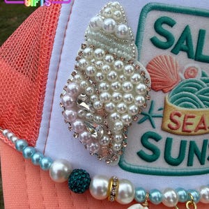 May include: A coral and white trucker hat with a beaded seashell design. The hat features a beaded strand with a seashell and starfish charm. The text "SALT SEA SUN" is embroidered on the hat.