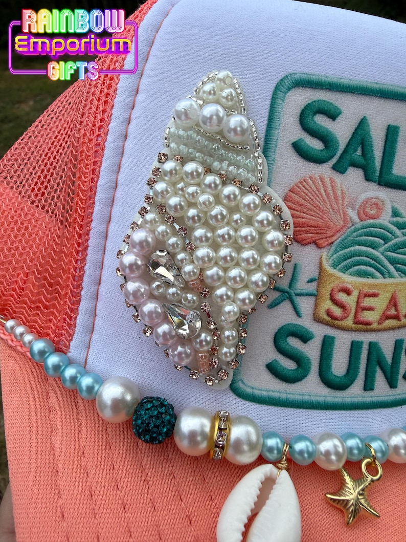 May include: A coral and white trucker hat with a beaded seashell design. The hat features a beaded strand with a seashell and starfish charm. The text "SALT SEA SUN" is embroidered on the hat.