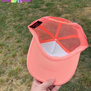 May include: Coral-colored trucker hat with a white front panel and mesh back. The hat features a curved brim and a snapback closure. The interior has a white sweatband and a tag that reads "OTTO". The text "RAINBOW Emporium GIFTS" is visible.