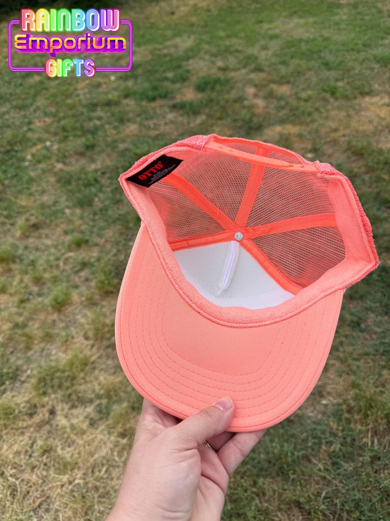 May include: Coral-colored trucker hat with a white front panel and mesh back. The hat features a curved brim and a snapback closure. The interior has a white sweatband and a tag that reads "OTTO". The text "RAINBOW Emporium GIFTS" is visible.