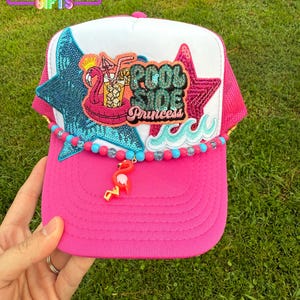 May include: Pink trucker hat with a white front panel, featuring a sequined star and a patch with a flamingo floatie and the words "Pool Side Princess". A beaded strand and flamingo charm hang from the brim.