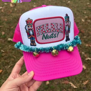 May include: A bright pink trucker hat with a white front panel featuring a "FALALA Nuts!" design and nutcracker illustrations. The brim is pink, and the hat is adorned with a teal garland and gold bells. The hat has the text "Rainbow Emporium Gifts" on the top left.