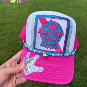 May include: A pink and white trucker hat with the text "Dive Bar Diva" on the front. The hat has a sequined crown and a beaded accent. The brim is bright pink, and the top is white with a pink mesh back.
