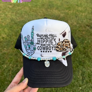 May include: Black and white trucker hat with the text "Raising Hell with the Hippies and the Cowboys." The hat features a turquoise beaded chain, a skeleton hand, and a leopard print cowboy hat patch.