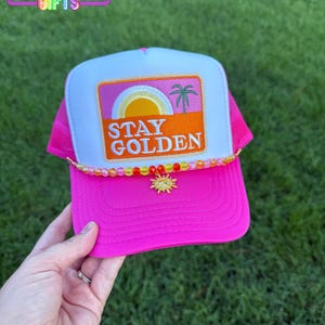 May include: A bright pink trucker hat with a white front panel featuring a sunset graphic and the words "STAY GOLDEN." The hat has a beaded brim and a sun charm. The text "RAINBOW Emporium GIFTS" is at the top.