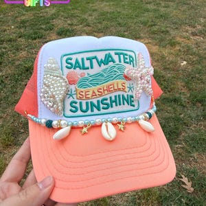 May include: A coral and white trucker hat with the text "Saltwater, Seashells, Sunshine". The hat is decorated with seashells, pearls, and a starfish. The brim is coral, and the mesh is orange.