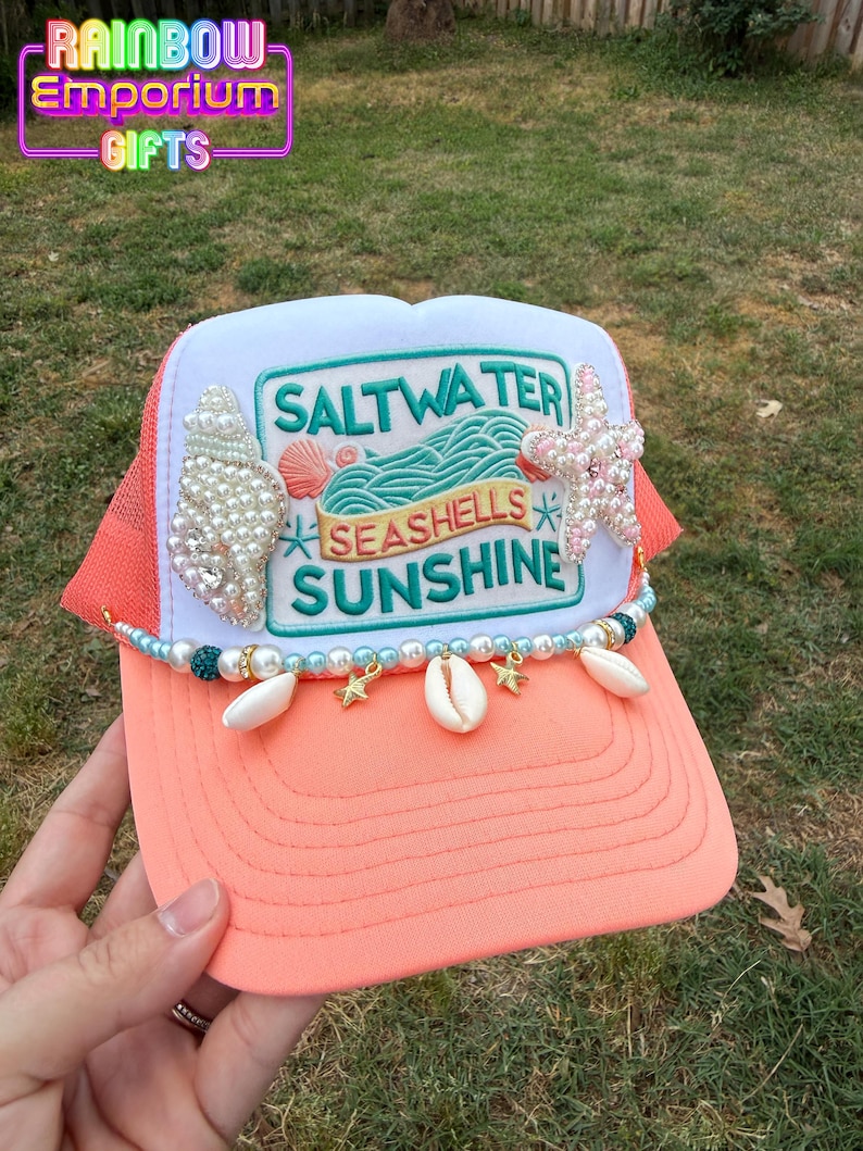 May include: A coral and white trucker hat with the text "Saltwater, Seashells, Sunshine". The hat is decorated with seashells, pearls, and a starfish. The brim is coral, and the mesh is orange.