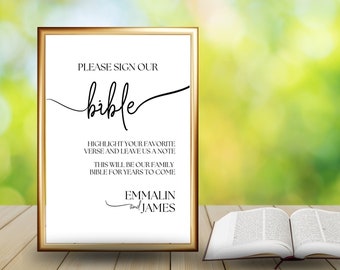 May include: A white sign with a gold frame, on a wooden table, with the text "PLEASE SIGN OUR bible HIGHLIGHT YOUR FAVORITE VERSE AND LEAVE US A NOTE THIS WILL BE OUR FAMILY BIBLE FOR YEARS TO COME EMMALIN and JAMES".