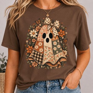 Peut inclure: Un t-shirt marron à manches courtes avec un motif central représentant un fantôme composé de divers tissus en patchwork, incluant des marguerites, des carreaux et des imprimés floraux. Le fantôme porte un nœud à pois et est entouré de fleurs.
