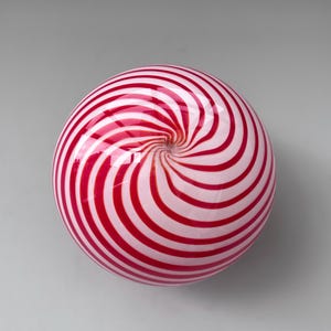 Op de afbeelding: Een ronde, glazen decoratieve bol met een rood-wit spiraalpatroon. Het glanzende oppervlak reflecteert het licht en benadrukt het wervelende patroon. Het object is waarschijnlijk mondgeblazen en heeft een gladde, gepolijste afwerking.