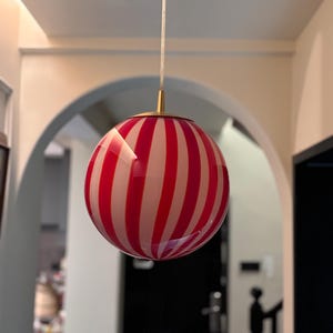 May include: A round, striped pendant light fixture with alternating red and white vertical stripes. The light hangs from a thin, white cord and has a gold-colored metal cap. The light fixture is a decorative element for interior spaces.