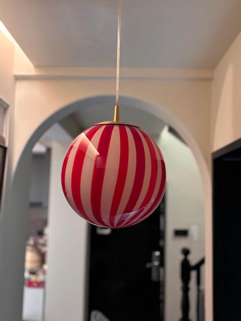 May include: A round, striped pendant light fixture with alternating red and white vertical stripes. The light hangs from a thin, white cord and has a gold-colored metal cap. The light fixture is a decorative element for interior spaces.