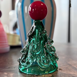 Puede incluir: Un adorno de árbol de Navidad de vidrio verde con una bola de vidrio roja en la parte superior. El árbol está decorado con espirales y tiene un acabado brillante.