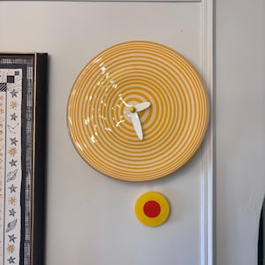 May include: A yellow and white striped wall clock with white hands. A small yellow circle with a red center is below the clock. The clock is mounted on a white wall.
