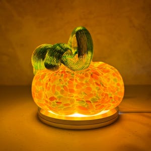 Può includere: Una scultura di zucca in vetro con un corpo maculato arancione, giallo e bianco e un gambo ricurvo verde traslucido. La zucca poggia su una base circolare bianca illuminata che proietta un bagliore caldo attraverso il materiale in vetro.
