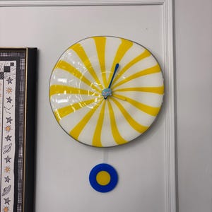 Puede incluir: Un reloj de pared caprichoso con una esfera a rayas blancas y amarillas, manecillas azules y una pieza de acento azul y amarillo debajo. El reloj está montado en una pared blanca. El diseño del reloj recuerda a una piruleta.