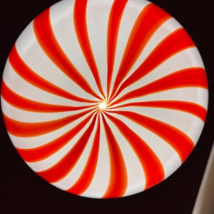 May include: A round, white plate with a red spiral design. The red stripes radiate from the center, creating a candy cane-like pattern. The plate is set against a dark background.