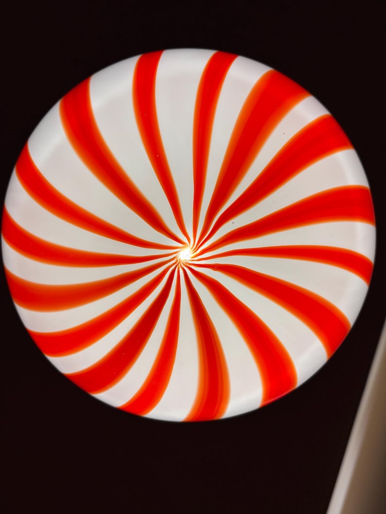 May include: A round, white plate with a red spiral design. The red stripes radiate from the center, creating a candy cane-like pattern. The plate is set against a dark background.