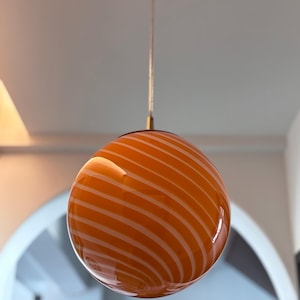 May include: Orange glass pendant light with white spiral stripes. The light fixture has a round shape and is suspended from a brass-colored fixture. The light is hanging from the ceiling.