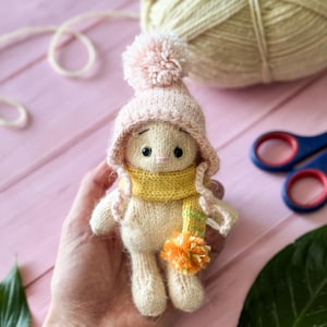 May include: A knitted toy bunny wearing a pink hat with a pom-pom and a yellow scarf with orange stripes. The bunny is sitting in a hand, with yarn and scissors in the background.
