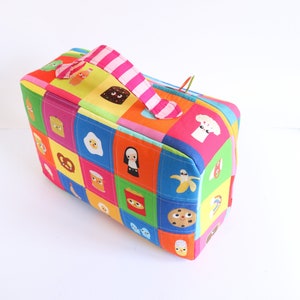 Puede incluir: Una caja de almacenamiento de tela colorida con un asa a rayas rosa y blanca. La caja est&aacute; decorada con un dise&ntilde;o de patchwork de varios personajes y objetos de dibujos animados, incluyendo un pl&aacute;tano, una galleta y un pretzel.