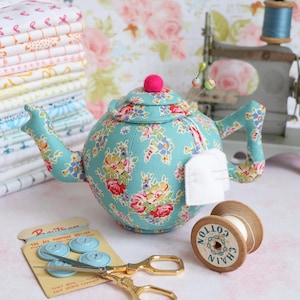 May include: A teal blue teapot pincushion with a floral pattern and a pink pom-pom on top. The teapot is filled with pins and has a white tea bag hanging from the spout. There is a spool of white thread, a pair of gold scissors, and a card of blue buttons on a pink and white floral background.