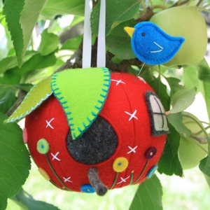 May include: A red felt apple birdhouse with a green felt leaf on top. The birdhouse has a small window and a round opening for birds to enter. There are small felt flowers and a blue felt bird on a pin attached to the birdhouse.