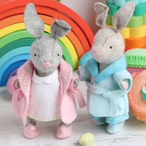 May include: Two grey felt bunny toys wearing different outfits. One bunny is wearing a pink coat with a white dress and pink shoes. The other bunny is wearing a blue robe with blue trousers and blue shoes. The bunnies are standing on a white wooden surface with green and yellow Easter eggs.