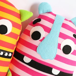 May include: Two plush toys with striped fabric and friendly faces. One is yellow and red with green horns, the other is pink and black with a blue nose.