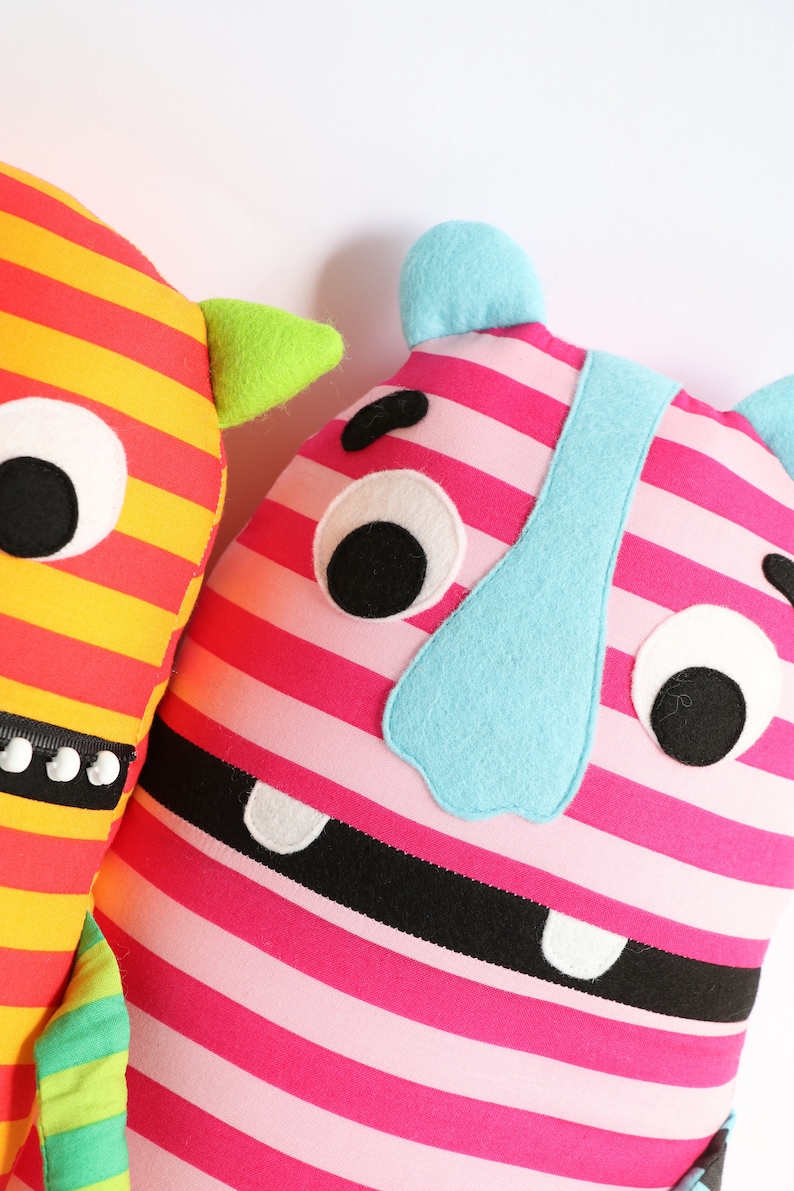May include: Two plush toys with striped fabric and friendly faces. One is yellow and red with green horns, the other is pink and black with a blue nose.