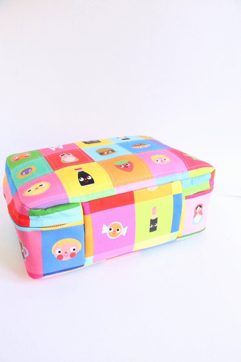 Puede incluir: Una bolsa rectangular con cremallera de colores con un dise&ntilde;o de patchwork que presenta varios personajes y objetos de dibujos animados. La bolsa est&aacute; hecha de tela y tiene un cierre de cremallera.