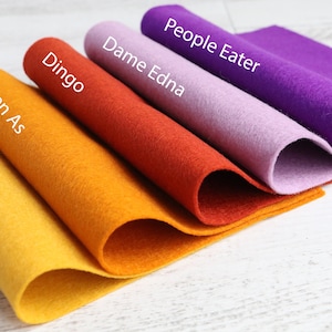 Puede incluir: Cinco telas de fieltro de diferentes colores, dobladas y apiladas sobre una superficie de madera blanca. Los colores del fieltro son amarillo, naranja, rojo, morado y morado claro. El fieltro est&aacute; etiquetado con los nombres "Gaytime", "Keen As", "Dingo", "Dame Edna" y "People Eater".
