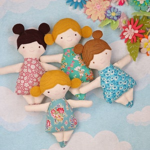 May include: Four handmade fabric dolls with different floral patterned dresses and brown or blonde hair. The dolls are arranged on a light blue background with white clouds.