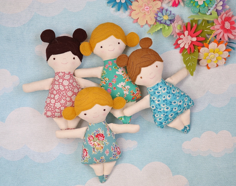 May include: Four handmade fabric dolls with different floral patterned dresses and brown or blonde hair. The dolls are arranged on a light blue background with white clouds.