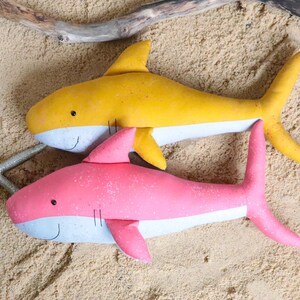 May include: Two plush shark toys on a sandy background. One shark is yellow with a white belly and the other is pink with a white belly.