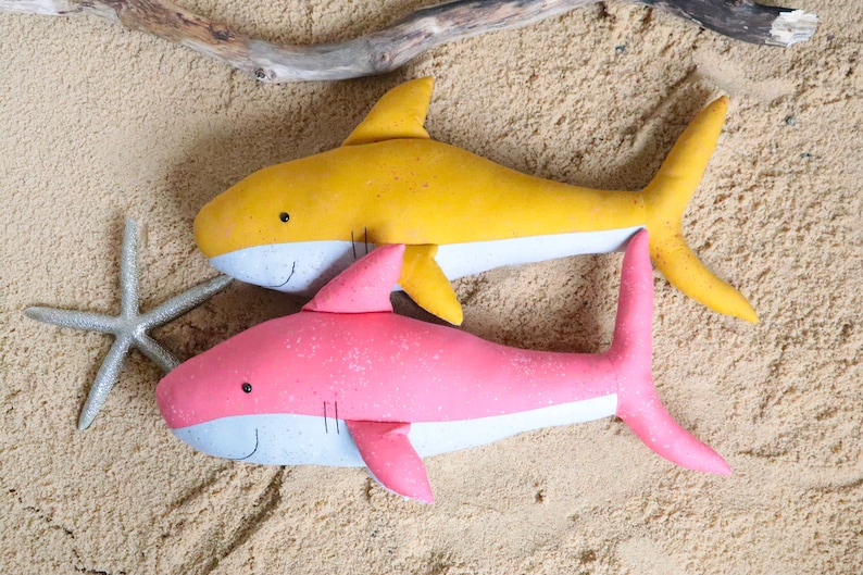 May include: Two plush shark toys on a sandy background. One shark is yellow with a white belly and the other is pink with a white belly.