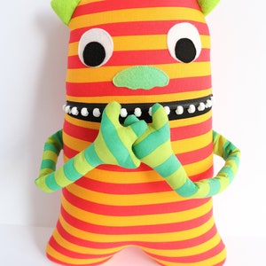 May include: A striped plush toy monster with red and yellow stripes, green arms, and a zipper mouth. The monster has large black eyes and a green nose.