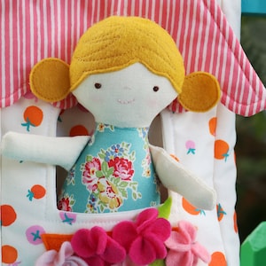 May include: A white fabric dollhouse with a pink and white striped roof. A doll with yellow hair, a blue floral dress, and white arms is peeking out of the window. The dollhouse has a pocket with pink and purple felt flowers.