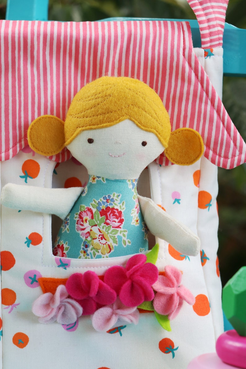 May include: A white fabric dollhouse with a pink and white striped roof. A doll with yellow hair, a blue floral dress, and white arms is peeking out of the window. The dollhouse has a pocket with pink and purple felt flowers.