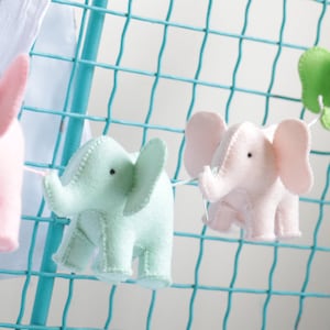 May include: A garland of felt elephant ornaments in pink and green hanging on a turquoise wire mesh background.