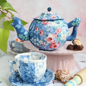 May include: A fabric teapot and matching teacup with saucer, both featuring a blue floral pattern with pink and white accents. The teapot has a lid with a small knob, and the teacup sits on a matching saucer. The set is made of quilted fabric.