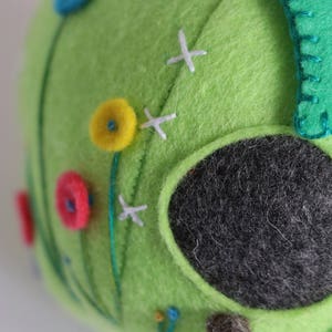 May include: A green felt birdhouse with a round opening, decorated with colorful felt flowers and white stitching. The birdhouse has a brown felt branch extending from the bottom.