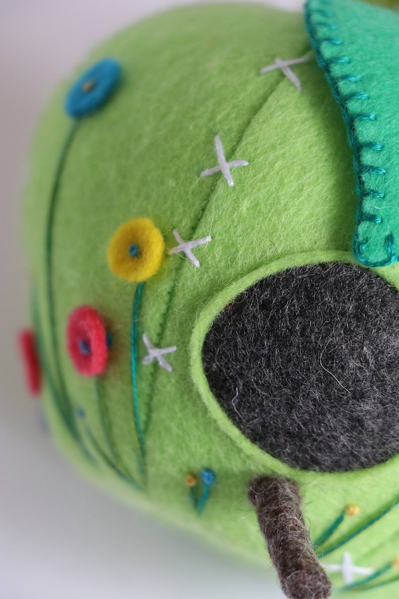 May include: A green felt birdhouse with a round opening, decorated with colorful felt flowers and white stitching. The birdhouse has a brown felt branch extending from the bottom.