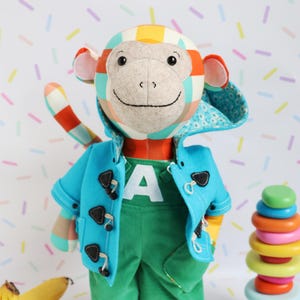 May include: A plush monkey doll with a colorful patterned head and tail, wearing a blue coat, green pants, and a shirt with a large white "A". The monkey has a smiling face and is next to a stack of colorful rings.