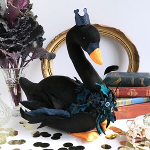 Può includere: Un cigno nero di peluche con becco e zampe arancioni, ornato da una corona blu e decorazioni floreali. Il cigno è decorato con piume e fiori verde acqua e neri. È un oggetto decorativo.