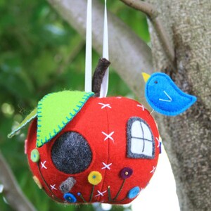 May include: A red felt apple birdhouse with a green felt roof, a white felt window, and a blue felt bird perched on the side. The birdhouse is decorated with colorful felt flowers and stitching.