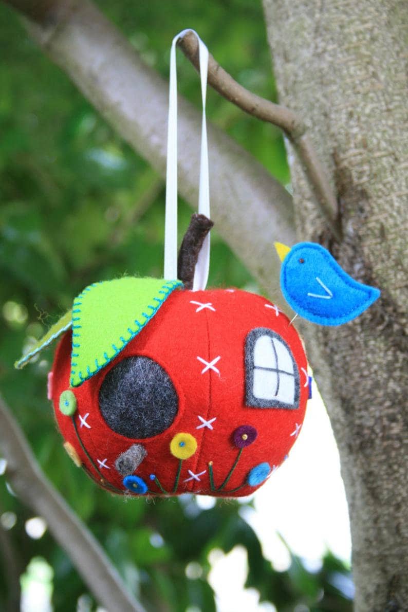 May include: A red felt apple birdhouse with a green felt roof, a white felt window, and a blue felt bird perched on the side. The birdhouse is decorated with colorful felt flowers and stitching.