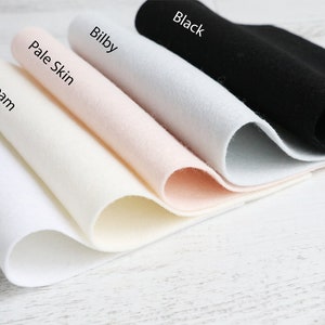 Puede incluir: Cinco muestras de tela de fieltro en diferentes colores: blanco, crema, piel p&aacute;lida, bilby y negro. Las muestras est&aacute;n dobladas y dispuestas en una fila de la m&aacute;s clara a la m&aacute;s oscura.