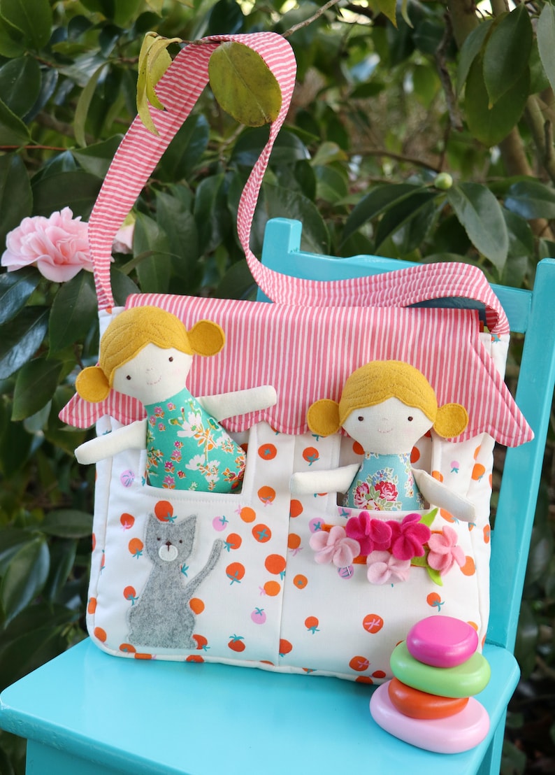 May include: A white fabric bag with pink and white stripes, featuring two pockets with felt dolls and a gray felt cat. The bag has a pink strap and is sitting on a blue chair.